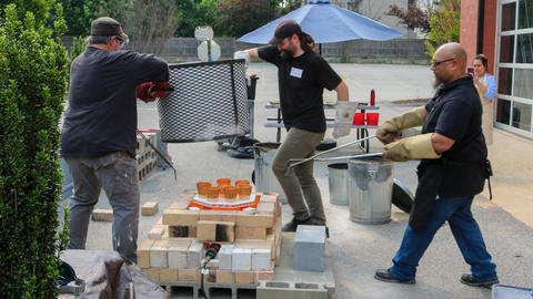 two people lifting a kiln cylinder and a third using tongs to pluck red-hot pots out of it during an outdoor raku firing 