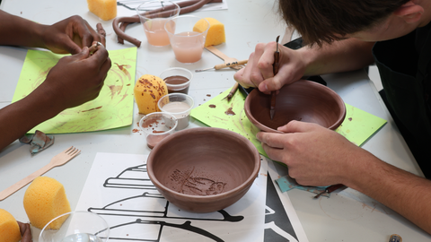 hands working a decoration onto a red clay bowl, on a table strewn with diagrams of historic forms