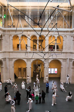 Aerial view of ON DISPLAY HARVARD 2023 in Harvard Art Museums' Calderwood Courtyard. Performers in all white clothing and paper wearables are posed. A white sculpture is in the center. Members of the public, comprising adults and children, are looking up close.