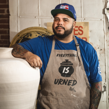 a person stands with their elbow propped on a human-size unglazed clay urn. They are wearing a baseball cap and an apron with the words, 