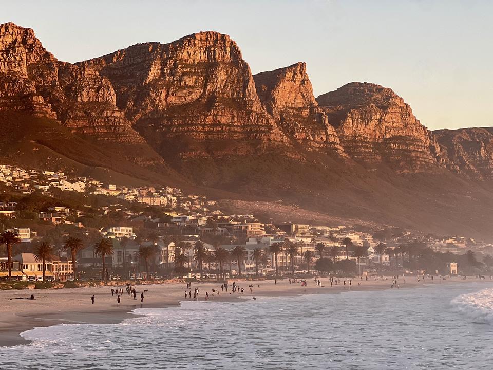 Photograph of a beach town beneath steep red cliffs at sunset, with waves breaking on the shore.