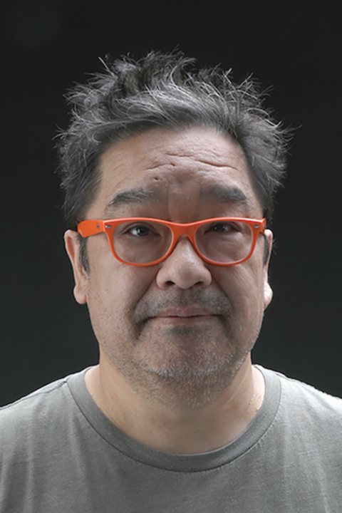 Shoji Satake looks at the camera, wearing orange-framed glasses and a gray crewneck shirt