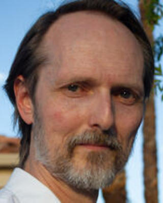 Close headshot of Aaron Jensen, a White man with a gray short beard wearing a white collared shirt. Behind him is a blue sky and palm tree.