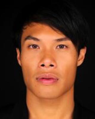 A close-up headshot of an Asian American man looking intensely at the camera. He has short black hair that falls over his forehead. Black backround.