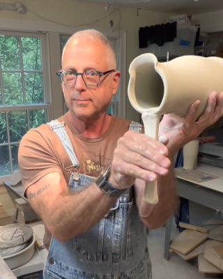 A potter holding a freshly thrown clay vessel while in the act of pulling a handle in wet clay directly off of the vessel