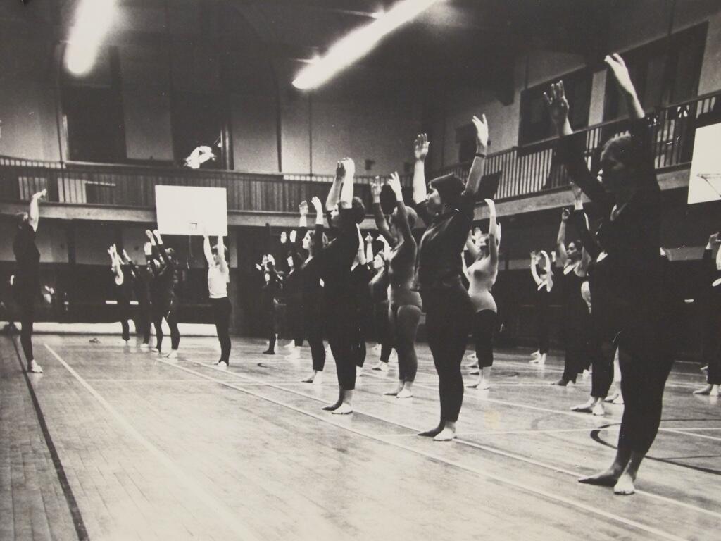 A black and white photo of rows of student dancers in the former Radcliffe Gym during a dance class. All the dancers are standing with their feet turned out and their arms up over their heads. A teacher is demonstrating at the front of the class. Photo circa 1970s and courtesy of Harvard Dance Center. 