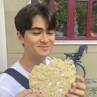 Chase Melton stands holding a crispy, circular-shaped food item. He is smiling down at it, surrounded by classic architecture and windows behind him