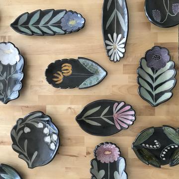Variety of glazed plates by artist Ruth Easterbrook with floral motifs. Most black with pastel colors.