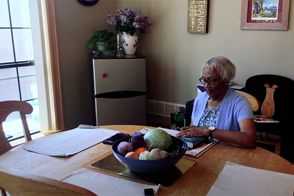 Screenshot from video. A woman sits at her dining room table, looking at a closed book.