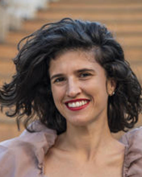 close-up of Laura Sanchez, a light skinned woman smiling into the camera. She has wavy brown hair neck-length, and red lipstick. Wearing a mauve dress.