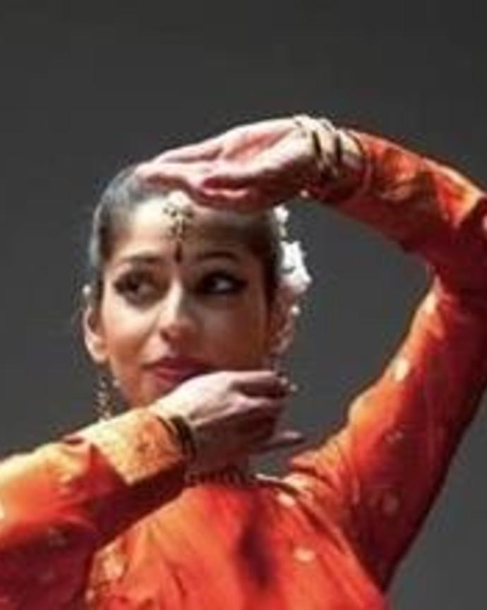 A close up of Anjali Nath dancing; she wears an orange tunic; her arms framing her face.